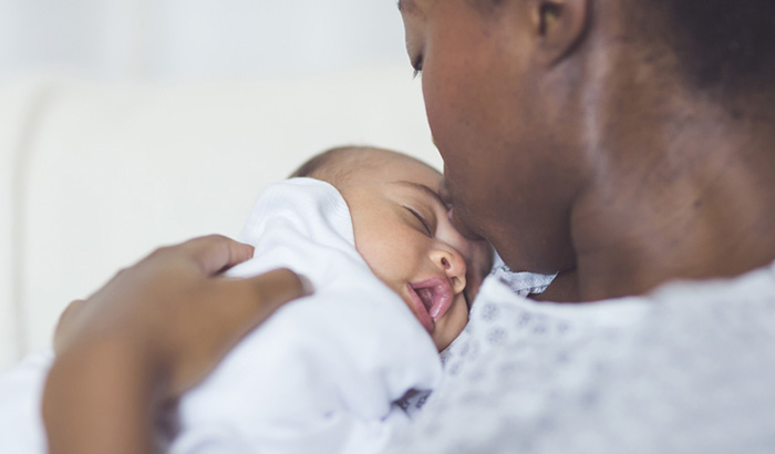 A beautiful young African American mother in a hospital gown gently holds her infant in her arms and smiles down at her. The swaddled baby's eyes are tightly closed.